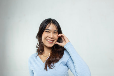 Beautiful young Asian woman with tan skin, toothy style smile with hand on her cheek, face and skin care concept, smooth blue top, white studio backgroundの写真素材