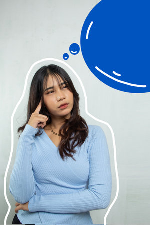A girl standing thinking about something in her head with her index finger, she is wearing a blue casual suit and a blue talk box beside her.の写真素材