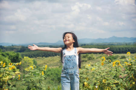 Pretty kid children asia girl in blue jean white shirt resting in sunflower field mountains background, arms wide open at natureの写真素材