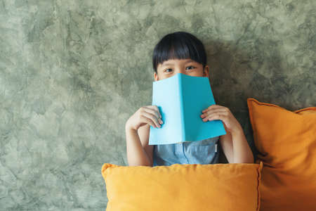 Photo of little asia girl sitting witn orange pillow and covered her face with blue book at coffee cafe shop. Looking camera covering face with book.の写真素材