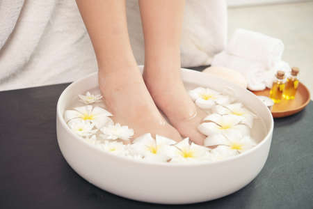 closeup view of woman soaking her feet in dish with water and flowers on wooden floor. Spa treatment and product for female feet and hand spa. white flowers in ceramic bowl.の写真素材