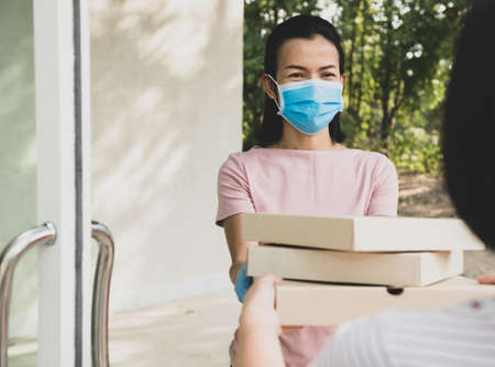 delivery service in t-shirt, in protective mask and gloves giving food order, holding three pizza boxes in front house, woman accepting delivery of boxes from delivery man during COVID-19 outbreak.の写真素材