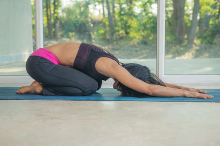 Young attractive woman practicing yoga exercise at home, working out, wearing sportswear, pants and top, indoor full length, Balasana, Child Pose, Ardha-Kurmasana. doing forward bend yoga on blue matの写真素材