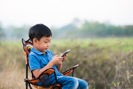 A boy looking at mobile phone. Sitting on a picnic chair in garden park.の写真素材