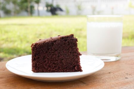 Chocolate cake on white plate. Put on wood table Selected focus on front. A glass of milk and garden  view are background.の写真素材