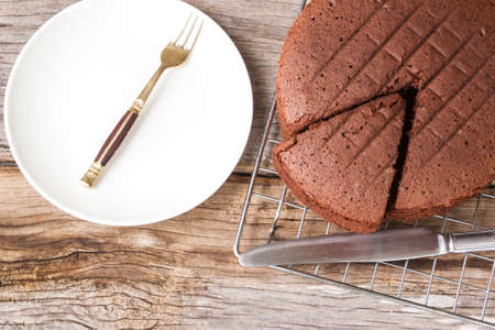 Chocolate cake on baking rack with knife. And fork on white plate. Over wooden table. Top view.