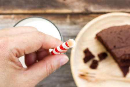 Woman's hand holding straw in a glass of milk. With chocolate cake on wooden plate are background. Selected focus on straw.の写真素材