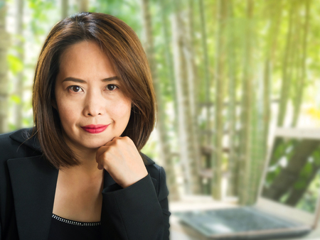 Asian business woman with blur green garden background. Smiling and positive emotion. Confident and looking to camera. Thai business woman were black suit and black singlet. Short hair and black eye with red lip.の写真素材