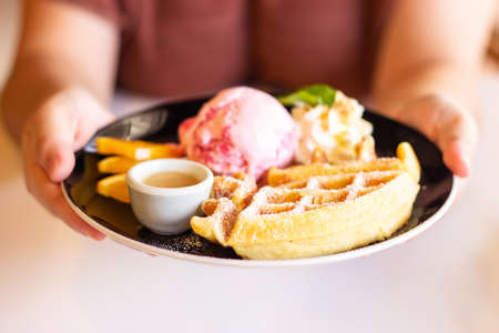 Woman hand holding plate of homemade waffle served with strawberry ice cream and fresh fruit. Sweet dessert. Pastel color tone.の写真素材