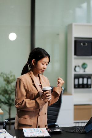 Happy Asian businesswoman working for her project and drinking coffee cup while working, sitting at the office desk.の写真素材
