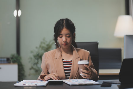 Happy Asian businesswoman working for her project and drinking coffee cup while working, sitting at the office desk.の写真素材