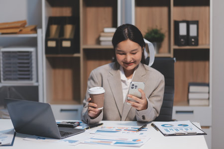 Positive asian young female accountant using a smartphone at her office desk. Woman relaxing on her mobile application.の写真素材
