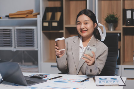 Positive asian young female accountant using a smartphone at her office desk. Woman relaxing on her mobile application.の写真素材