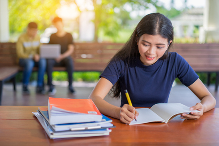 Female student taking notes from a book at library, Young asian woman sitting at table doing assignments in college libraryの写真素材