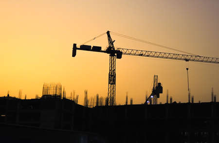 Silhouette of construction cranes and orange sky at dusk.