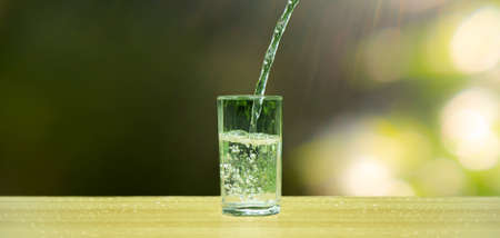 Pour water into a clear glass. Water flows into a clear cylindrical glass. Garden background,...