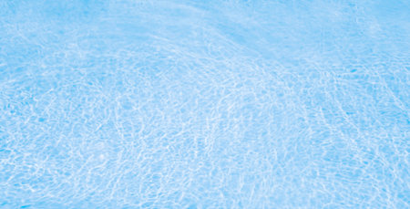 Rippling, clear blue water from swimming pool with light-colored bottom. Light from above creates...