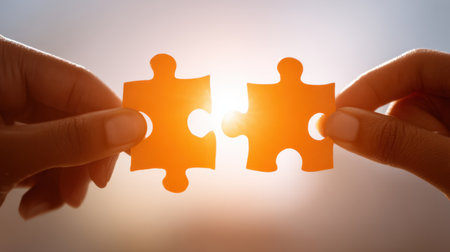 Two hands join vibrant orange puzzle pieces, symbolizing connection and collaboration. The warm background enhances the sense of unity and creativity in problem solving.の素材