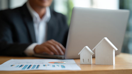 A business professional evaluates real estate data on a laptop with miniature house models present, embodying a modern office setting focused on investment and growth strategies.の素材