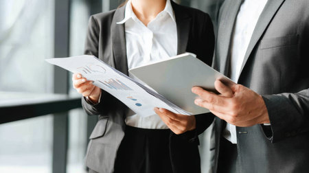 Two business professionals actively engage in a discussion while reviewing reports and a digital tablet, highlighting collaboration in a modern office setting.の素材