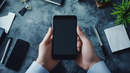 A close-up view of hands holding a smartphone with a blank screen, set on a stylish desk filled with office supplies, plants, and modern tools, ideal for business or tech themes.の素材