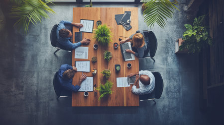 Aerial view of professionals engaged in an important meeting, working on laptops surrounded by greenery and documents in a modern office setting, fostering collaboration and creativity.の素材