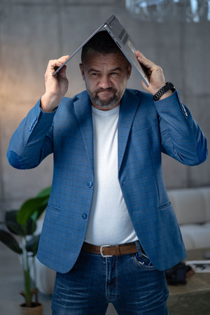 Bearded young confident guy stand behind office desk with laptop thinking working alone. handsome male in casual wear engaged in office work, planning strategy, deadlines. at workplaceの写真素材