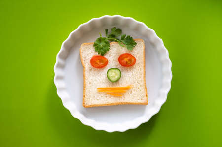 Fun food for kid, emotion face sandwich bread with vegetables white plate on green tableの写真素材