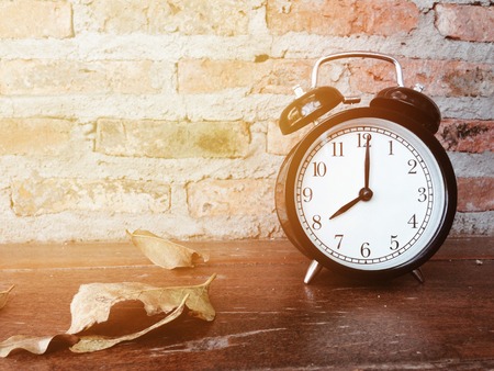 Retro black alarm clock with eight o'clock and dry leaves on wooden table and brick wall backgroundの写真素材