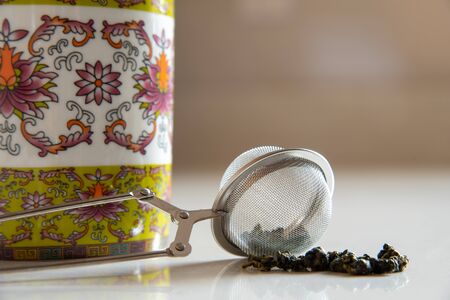 Selective focus of green tea pellets with a stainless steel ball infuser infront of an ornate mug