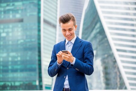 Handsome businessman in suit standing at the street with smart phone in hand. Modern skyscrapers on the background.の写真素材