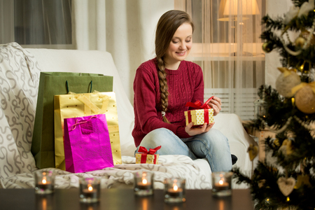 Happy young woman in red sweater sitting in the living room with decorated Christmas tree. Candles and christmas lights. Opening golden gift box with red ribbon. Happy holiday concept.の写真素材