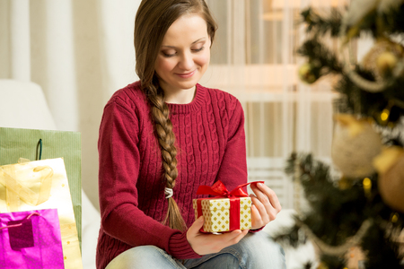 Happy young woman in red sweater sitting in the living room with decorated Christmas tree. Candles and christmas lights. Opening golden gift box with red ribbon. Happy holiday concept.の写真素材