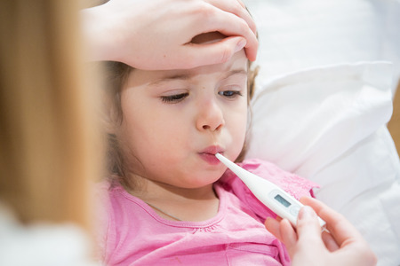 Mother measuring temperature of her ill kid. Sick child with high fever laying in bed and mother holding thermometer. Hand on forehead.の写真素材