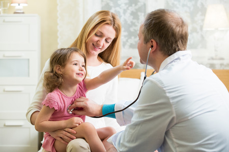 Male Pediatrician examining cute little girl with stethoscope. Kid happily smiling and pointing on doctor. Mother holding her kid. Doctor visit his patient at home.の写真素材