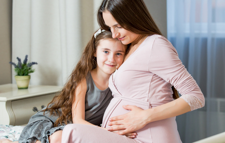 Portrait Of Affectionate family At Home. Pregnant healthy mother and daughter cuddling, kissing and looking at each other's eyes. Interior of cozy and stylish bedroom.の写真素材