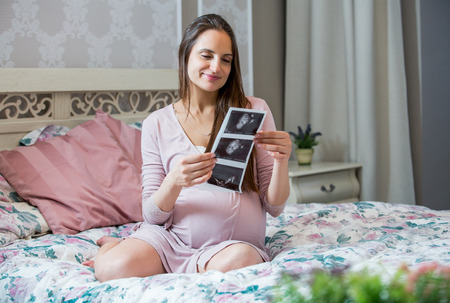 Beautiful healthy pregnant woman sitting on bed in cozy and stylish bedroom. Looking at ultrasound...