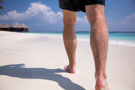 Man's bare feet walking at white sand beach. Picturesque landscape. Blue sky, coconut palms, white sand and azure water. Muscular legs walking on the beach. Leaving footprints on sand. Maldives.の写真素材