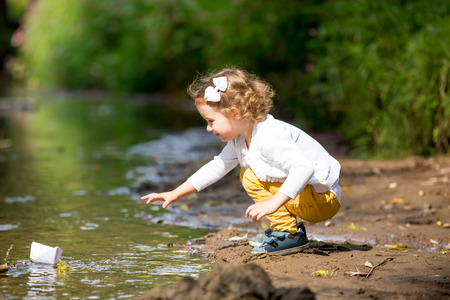 Cute little girl runs a paper boat in the stream in the park. Stretching her hand and reaching the little shipの写真素材