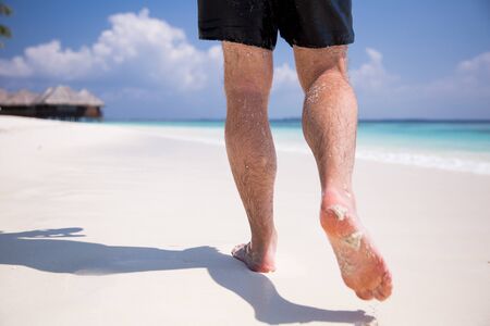 Man's bare feet walking at white sand beach. Picturesque landscape. Blue sky, coconut palms, white sand and azure water. Muscular legs walking on the beach. Leaving footprints on sand. Maldives.の写真素材