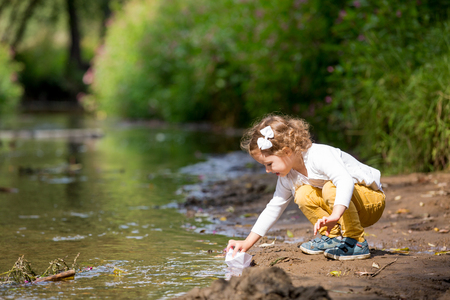 Cute little girl runs a paper boat in the stream in the park. Stretching her hand and reaching the little shipの写真素材