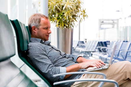 Casual business man working on laptop in airport hall. Man waiting his flight at airport terminal, sitting on chair and typing on the laptop.の写真素材