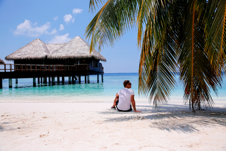 Man sitting under the coconut palm on tropical beach with white sand and azure water. Looking at the ocean, thinking in loneliness. Relaxing and enjoying beautiful view.の写真素材