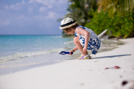 Sute little girl in beautiful dress and hat playing with sand on the tropical beach with white sand, azure blue sea and coconut palms. Paradise Landscape background.の写真素材