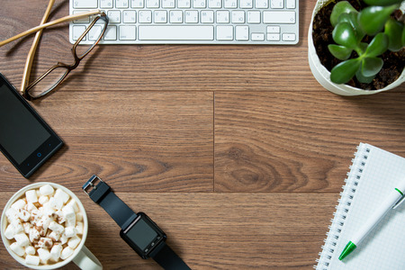 Business workplace from top, flay lay. Keyboard, smart watch, smart phone, glasses, paper notebook, hot chocolate with marshmallow on the dark wooden deskの写真素材