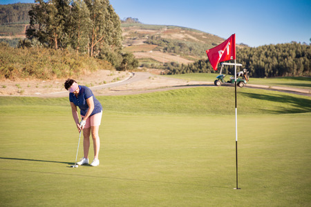 Mature woman playing golf with putter club on course. Golfer concentrating for putting on green. Beautiful sunny Landscape, green hills, blue sky. Portugal.の写真素材
