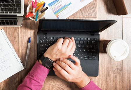 Business person working at office desk, typing and using laptop, top view flat lay. Checking smart watch on hand. Coffee cup, notepad, graphics around the workplace.の写真素材