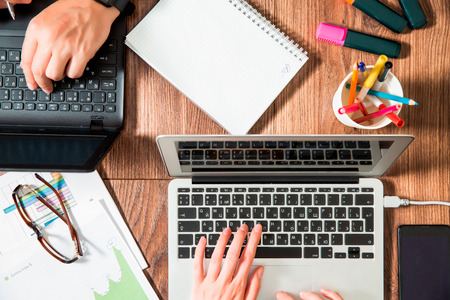 Two business persons working at office desk, typing and using laptop, top view, flat lay. Smart watch on hand and smart phone on the table. Coffee cup, glasses, folder, graphics around the workplace.の写真素材