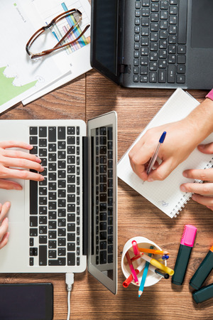 Two business persons working at office desk, typing, writing and using laptop, top view, flat lay. Smart phone on the table. Coffee cup, graphics, notepad and glasses around the workplace.の写真素材