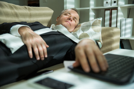 Businessman in suit lying on a couch with two cellphones and laptops, sleeping. Exhausted man relaxing in office early morning. Responsible executive working fell asleep.の写真素材